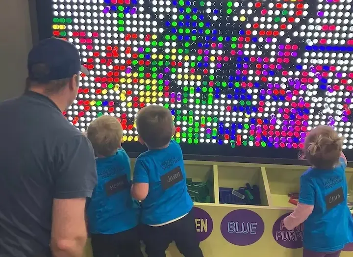 Field Trip to the Children's Museum. An adult male chaperone and three children with name tags on their backs playing with a colored light board.