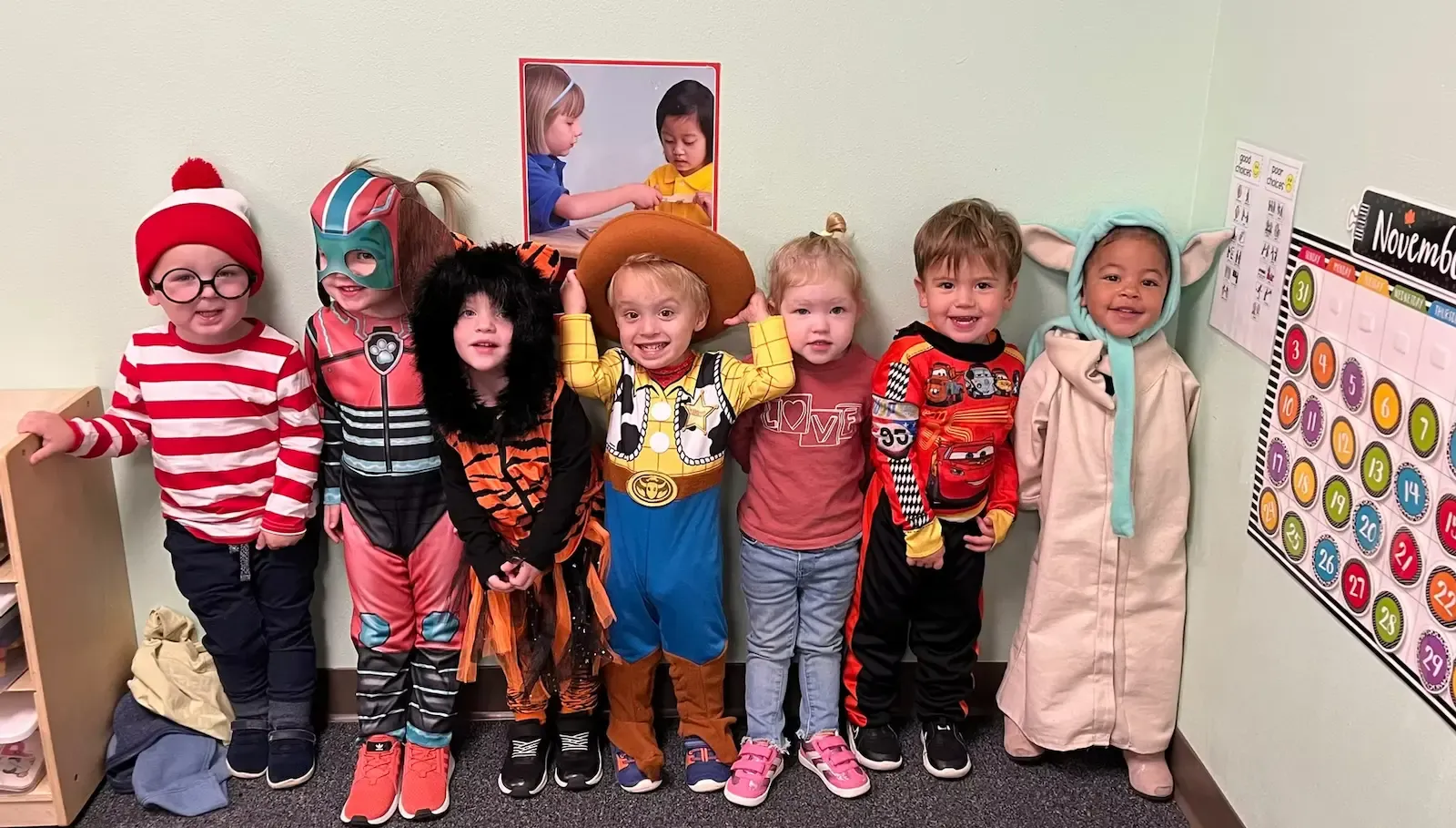7 children in a line against a wall, facing the camera. They are wearing halloween costumes and smiling.